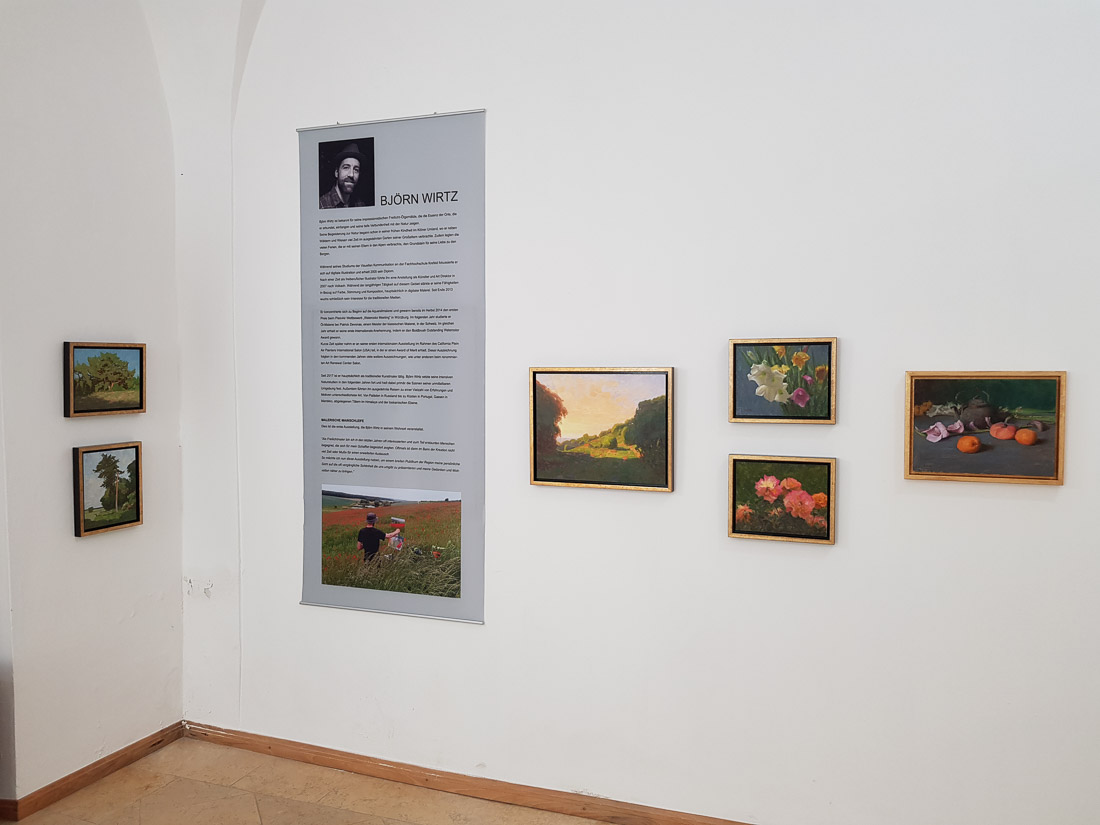 A corner of a room with white walls and paintings hanging next to a banner with information about the artist.
